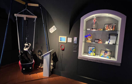 Das Bild zeigt einen Teil der Sonderaustellung Glück - Sonderausstellung Glück - Happiness im Museum der Stadt Villach. Auf der linken Seite hängt ein gleitschirm-sitz in den man sich reinsetzen kann. Über den Sessel hängt ein VR-Brille mit der man einen Gleitschirmflug miterleben kann. Auf der Rechten Seite befindet sich eine Vitrine mit Ausstellungsgegenständen. Die Wände sind in Schwarz gehalten. Der Sessel und die Vitrinen sind hell erleuchtet.
