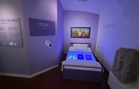 Das Bild zeigt einen Teil der Sonderaustellung Glück - Sonderausstellung Glück - Happiness im Museum der Stadt Villach. Auf den Bild ist ein Bett zu sehen auf denen Statistiken projiziert werden über das Familienleben der Kärntener. Oberhalb des Bettes hängt ein Bild von Jesus Christus. Der Raum ist in den Farben lila gehalten.