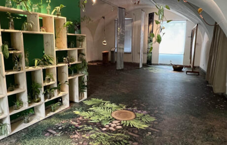 Das Bild zeigt einen Teil der Sonderaustellung Glück - Sonderausstellung Glück - Happiness im Museum der Stadt Villach. Auf den Bild erkennt man eine Übersicht eines Raumes der Sonderausstellung. Dieser Raum behandelt das Theme Glück in der Natur. Der Boden ist ein Fotoboden auf den ein Waldboden bedruckt ist. Links steht ein helles Holzregal mit verschiedenen Planzen. An der Decke hängen verschieden Kletterplanzen.